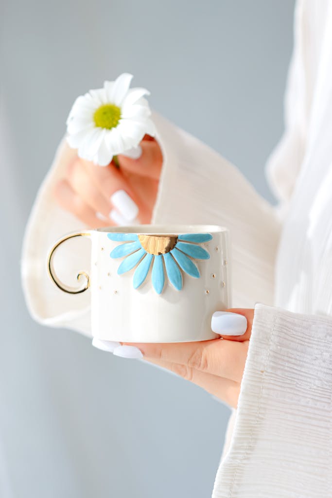 A woman holds a ceramic mug with floral design and a daisy, emphasizing elegance and simplicity.